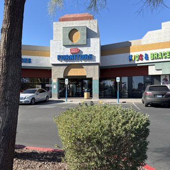 A strip mall storefront with multiple retail brands visible, including Furniture Store, Kids & Braces, and another store sign on the left. The building has a beige and gray facade with a circular window detail above the furniture entrance.